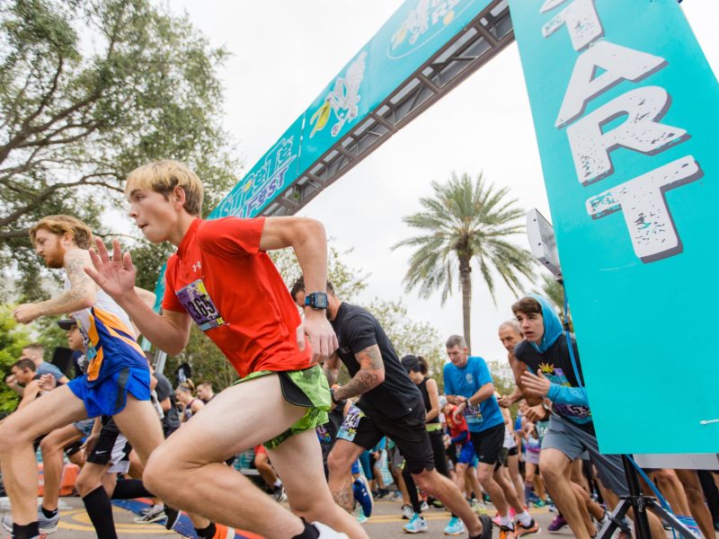 A low-angle action shot captures a group of runners as they burst forward from the starting line of a race. A large teal banner on the right reads "START" vertically in white letters. A young person in a red t-shirt and green shorts is in the foreground, mid-stride, surrounded by other competitors. A palm tree is visible in the background under an overcast sky.