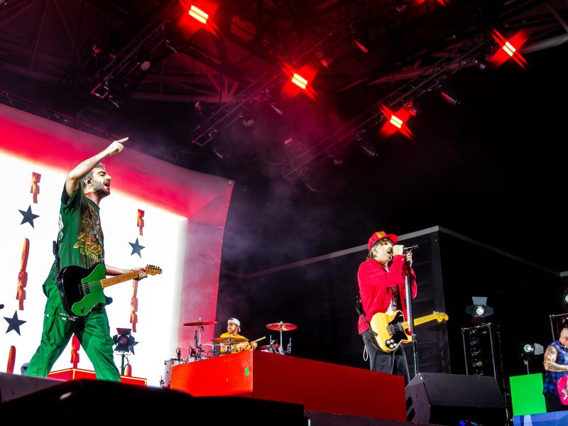 A wide shot of the band All Time Low performing on a dimly lit stage. Two guitarists stand on a raised platform in the foreground. On the left, a man with gray hair and a green shirt plays a black electric guitar, smiling toward the front. In the center, a man in a red jacket, red cap, and dark pants sings into a microphone while playing a light brown electric guitar. A third band member with a mohawk is visible on the right, also playing guitar. A drummer is partially visible in the lower left corner. The stage is dark, illuminated by red and white stage lights.