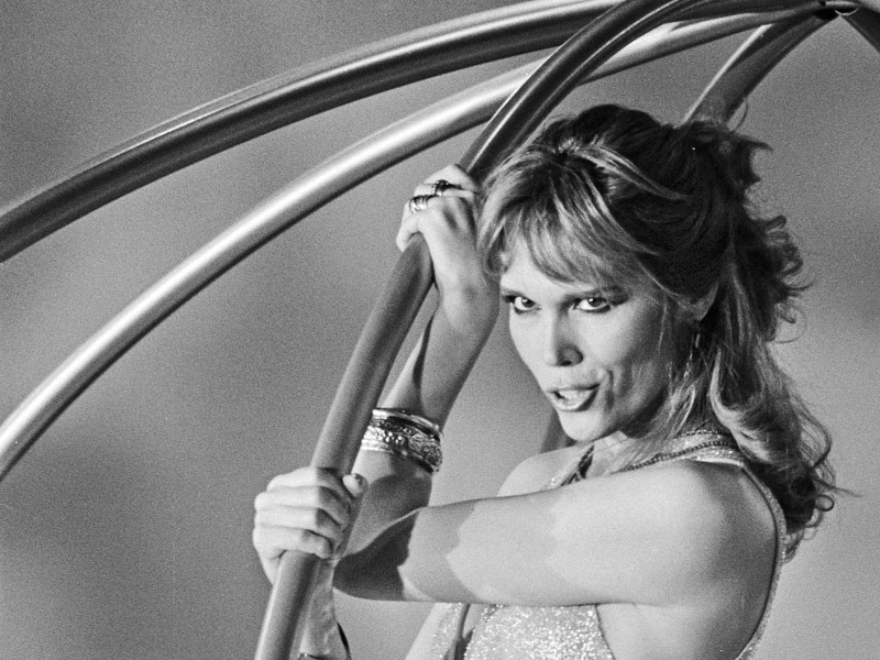 Black-and-white photo of Amanda Lear holding curved stage props, wearing a sparkling outfit and jewelry, with styled hair and a confident expression