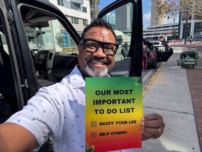 A smiling man wearing glasses and a white button-up shirt stands next to an open vehicle door and holds a sign that reads: "OUR MOST IMPORTANT TO DO LIST: ENJOY YOUR LIFE, HELP OTHERS" against a city street backdrop.