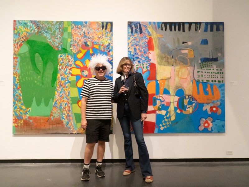 Two people stand in an art gallery between two large, colorful abstract paintings. One person wears a striped shirt and shorts with white spikey hair; the other wears a dark blazer, jeans, and sunglasses.