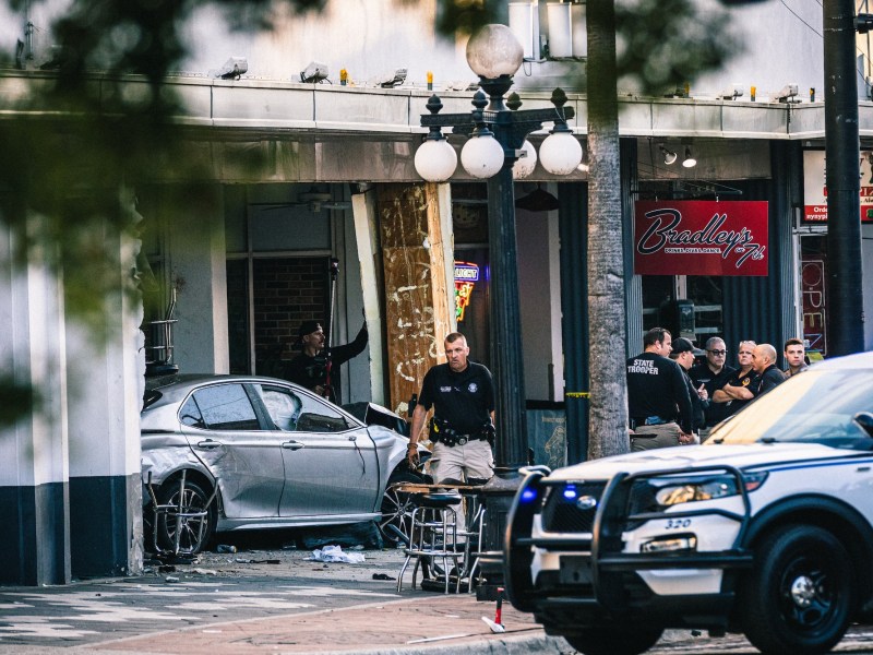 A silver car rests heavily damaged after crashing into the storefront of a business named 'Bradley's' in Ybor City. Police and State Troopers are gathered at the scene, with a patrol car visible in the foreground.