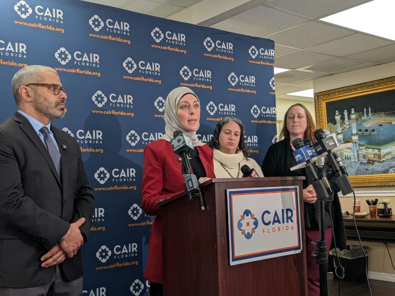 A woman wearing a red blazer and a hijab stands at a wooden podium speaking into several news microphones during a press conference. She is flanked by a man in a dark suit and glasses to her left, and two women standing to her right. The podium and the blue backdrop behind the group display the logo and website for "CAIR Florida." A framed picture of the Great Mosque of Mecca is visible on the wall in the background.