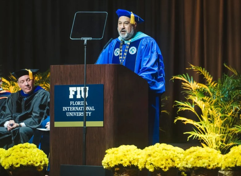 A man with a gray beard stands at a wooden podium delivering a speech, dressed in bright blue academic regalia with a matching mortarboard cap and a ceremonial chain of office. The podium displays the logo for "FIU Florida International University," and a transparent teleprompter screen stands in front of the speaker. To his left, another man in black academic robes sits listening. The foreground is lined with bright yellow flowers, and a palm plant is visible to the right against a dark curtain backdrop.