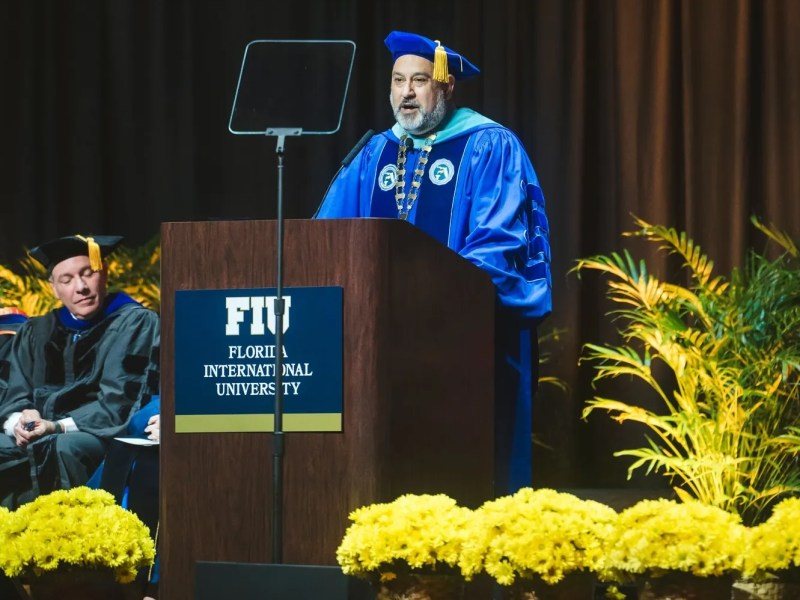 A man with a gray beard stands at a wooden podium delivering a speech, dressed in bright blue academic regalia with a matching mortarboard cap and a ceremonial chain of office. The podium displays the logo for "FIU Florida International University," and a transparent teleprompter screen stands in front of the speaker. To his left, another man in black academic robes sits listening. The foreground is lined with bright yellow flowers, and a palm plant is visible to the right against a dark curtain backdrop.