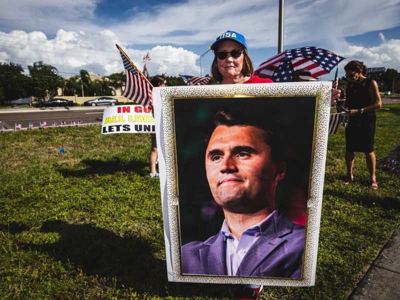 Low-angle shot of a woman wearing a blue "USA" hat holding a large, vertical, framed photo of Charlie Kirk and an American flag. She stands on a grassy area beside a paved road lined with small American flags. In the background, someone holds an American flag umbrella and there is a woman in a black dress.
