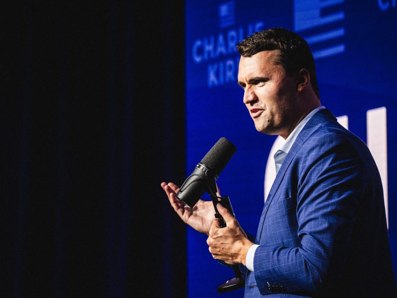 Speaker Charlie Kirk stands on a stage, wearing a blue patterned suit and white shirt, holding a microphone while speaking in profile against a blue backdrop.