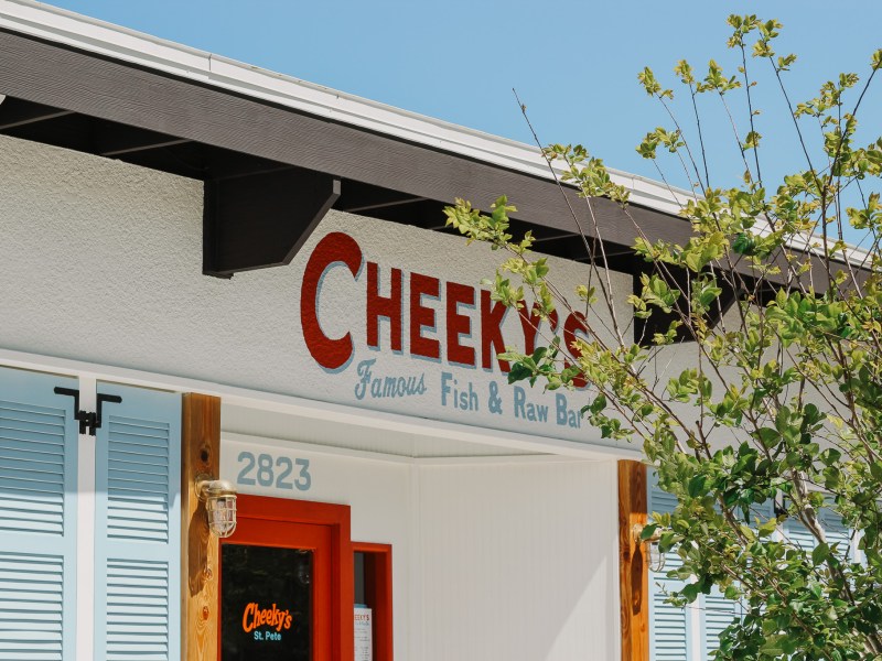A low-angle exterior shot of a white building with light blue shutters and a red door. Above the door, the business name "CHEEKY'S" is prominently displayed in a red, textured font, with "Famous Fish & Raw Bar" written beneath it in a lighter blue script. The number "2823" is visible to the left of the door. A black awning or beam runs across the top of the building facade. To the right, green foliage and tree branches are visible against a clear blue sky.
