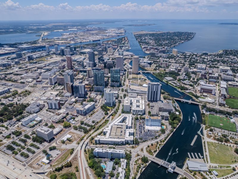 High aerial view of the downtown Tampa skyline, showing dense high-rise buildings along the Hillsborough River, which flows into Tampa Bay. Highways and urban development surround the central core.