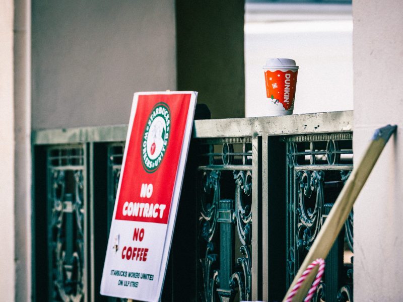 A Dunkin' disposable coffee cup sitting on a railing or ledge, with a blurred sign (mostly red and white) in the foreground to the left. The cup has a seasonal design, featuring a snowflake pattern and what looks like a tiny leaf or berry drawing.