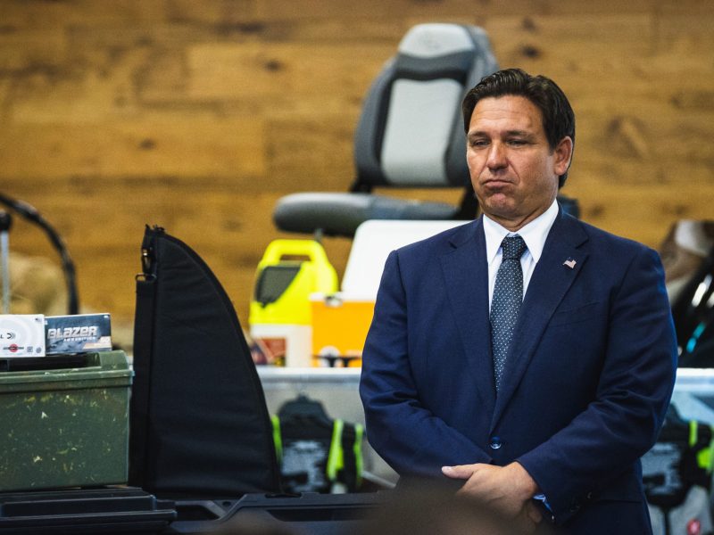 Governor Ron DeSantis, wearing a dark navy suit and tie, stands with a serious expression at an outdoor or indoor event. The background includes a wood-paneled wall and various items suggesting an outdoor theme, such as a boat seat, what appears to be a gun case or cover, and ammunition boxes.