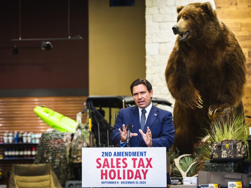 Governor Ron DeSantis speaks at a podium inside an outdoor supply store, with a large, taxidermied brown bear standing to his right. The podium sign announces a "2ND AMENDMENT SALES TAX HOLIDAY."
