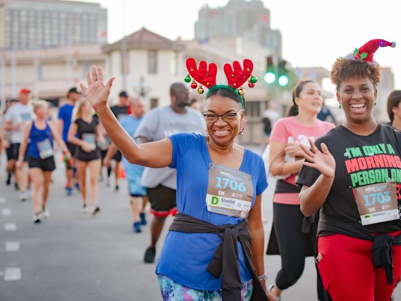 Two women smile and wave while participating in an outdoor race. The woman on the left wears a blue shirt, glasses, and a headband with reindeer antlers and ornaments, while the woman on the right wears a black shirt with the text "I'M ONLY A MORNING PERSON ON" and a Santa hat headband. Both are wearing race bibs for a "Hot Chocolate 15k/5k" event, with other runners visible in the blurred background.