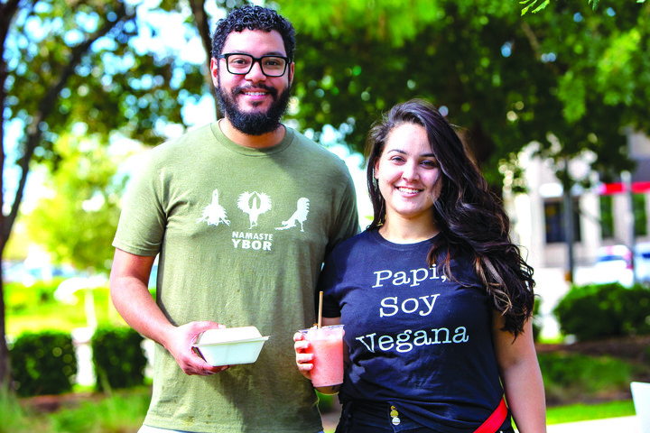 A waist-up, outdoor shot of a man and a woman smiling at the camera. The man on the left has a beard and glasses, wearing a green t-shirt that reads "NAMASTE YBOR," and holds a white food container. The woman on the right wears a black t-shirt with the text "Papi, Soy Vegana" and holds a pink smoothie. The background is brightly lit with blurred green trees.