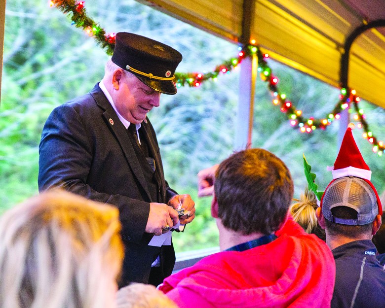 A smiling train conductor wearing a traditional black uniform and peaked cap stands in the aisle of a train car, punching a ticket for a passenger. The train is decorated for the holidays with green garland and multicolored Christmas lights hanging above the windows. In the foreground, other passengers watch, including one wearing a Santa hat headband.