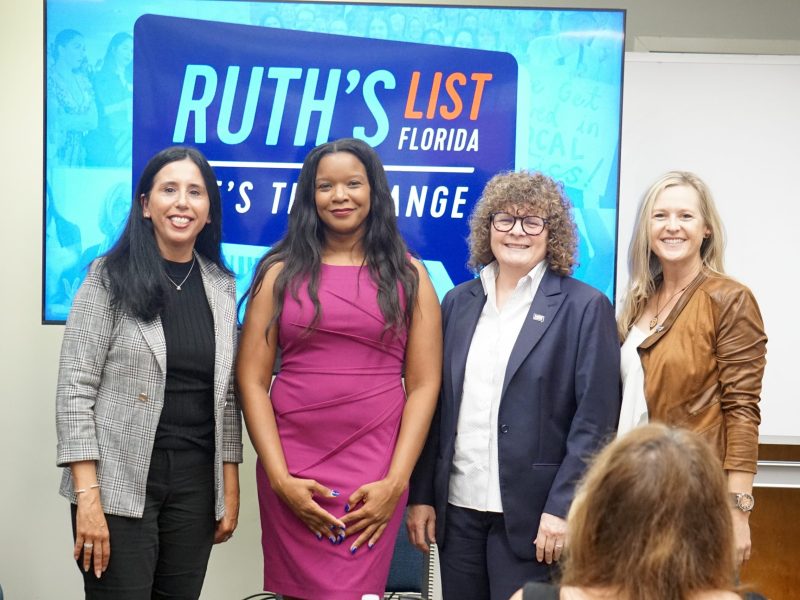 Four women stand side-by-side for a group photo in front of a screen displaying the "Ruth's List Florida" logo. The back of an audience member's head is visible in the foreground.