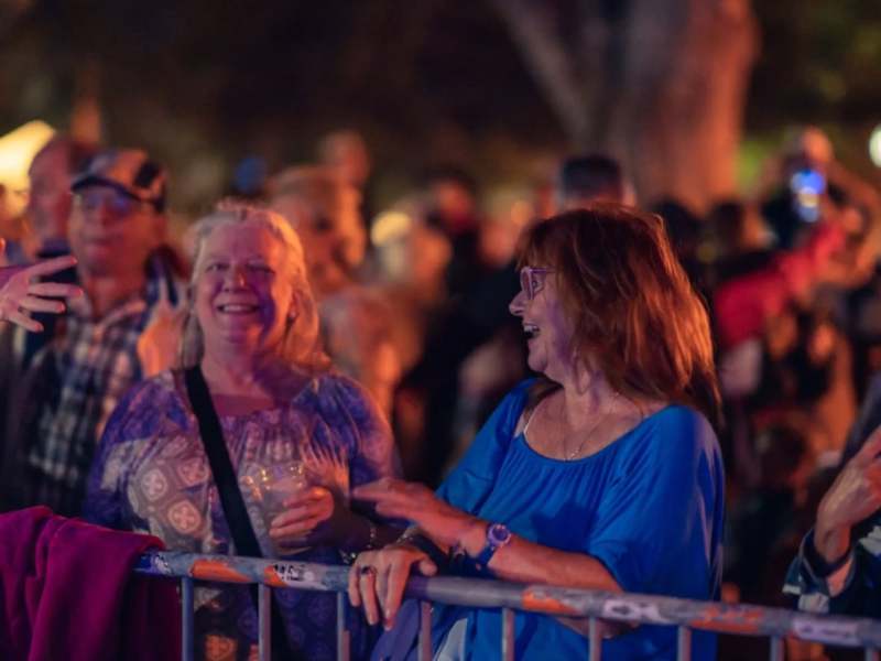 Two women stand behind a metal barrier, laughing and smiling at each other in a dense crowd during an evening music festival or event.