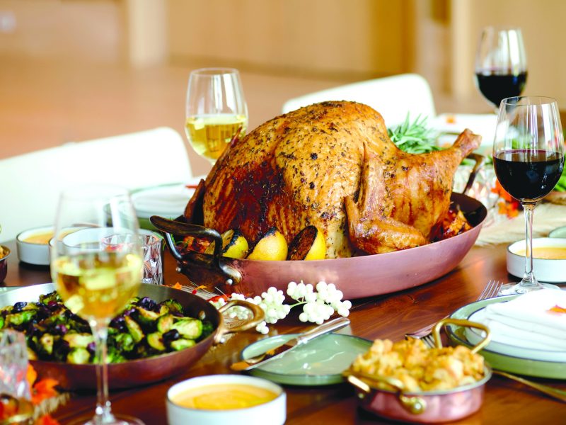 A close-up shot of a Thanksgiving dinner table, with a large, golden-brown roasted turkey in a copper pan as the centerpiece. The turkey is surrounded by side dishes, including Brussels sprouts and macaroni and cheese, and glasses of red and white wine are placed at various settings on the wooden table.