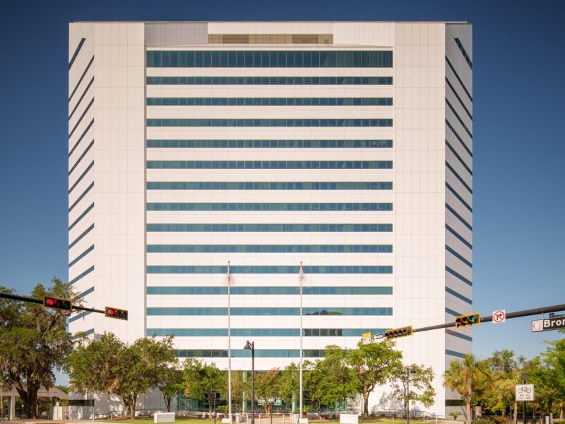 The Ralph D. Turlington Florida Education Center building, a tall white government structure with horizontal rows of windows, stands against a clear blue sky, viewed from a street intersection below.