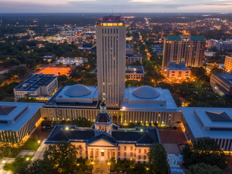 A bird's-eye view of the Florida State Capitol buildings. The classic, dome-topped Old Capitol is in the foreground, flanked by the modernist tall office tower and two shorter buildings with white domes. The city lights of downtown Tallahassee glow in the background.