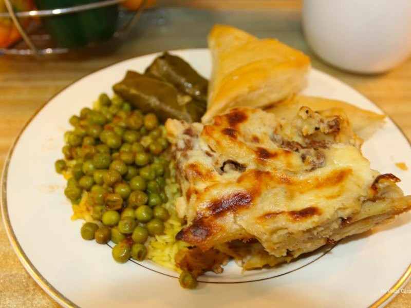 A white plate filled with various Greek foods, featuring a large, rectangular slice of pastitsio or moussaka with a browned, cheesy bechamel top. Alongside it are green peas, two stuffed grape leaves (dolmades), and a triangular piece of spanakopita.