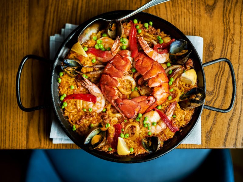 A top-down view captures a vibrant seafood paella served in a large black pan with two handles, set on a wooden table. The dish features yellow saffron rice mixed with green peas and red peppers, topped generously with mussels, clams, shrimp, calamari rings, and two large lobster tails in the center. A silver spoon rests on the edge of the pan, which is placed on a folded light blue napkin.