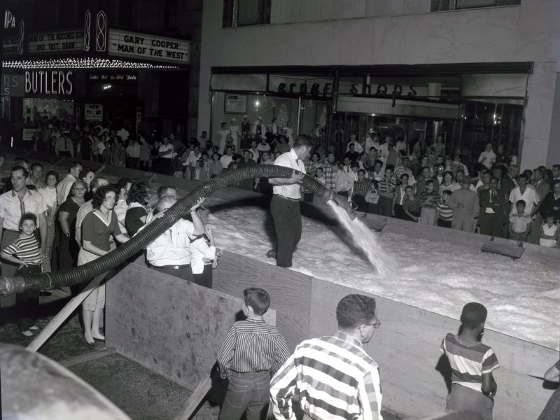 A black-and-white historical photograph depicts a man holding a large hose, spraying a pile of snow or ice into a wooden enclosure on a city street at night. A large crowd of adults and children stands behind the enclosure to watch the event. In the background, illuminated storefronts and theater marquees are visible, including one advertising the Gary Cooper film "Man of the West" and another reading "Butlers".