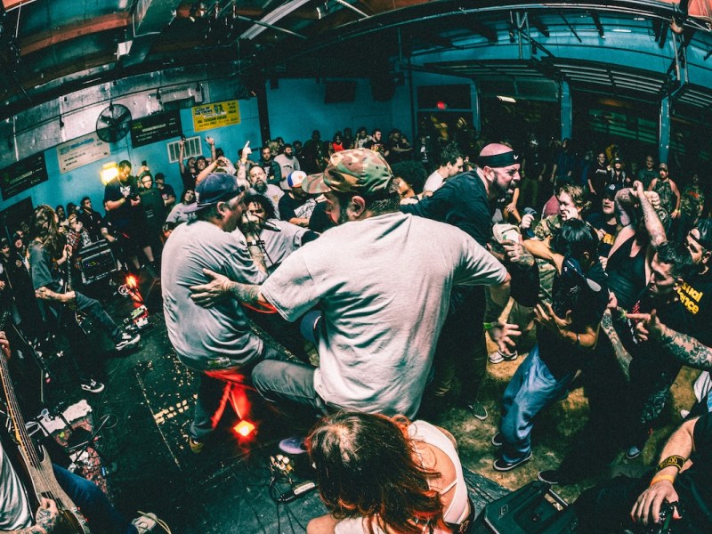 Wide-angle view of a densely packed, high-energy punk or hardcore concert, with audience members standing on the stage and leaning into the crowd, creating a mosh pit atmosphere.