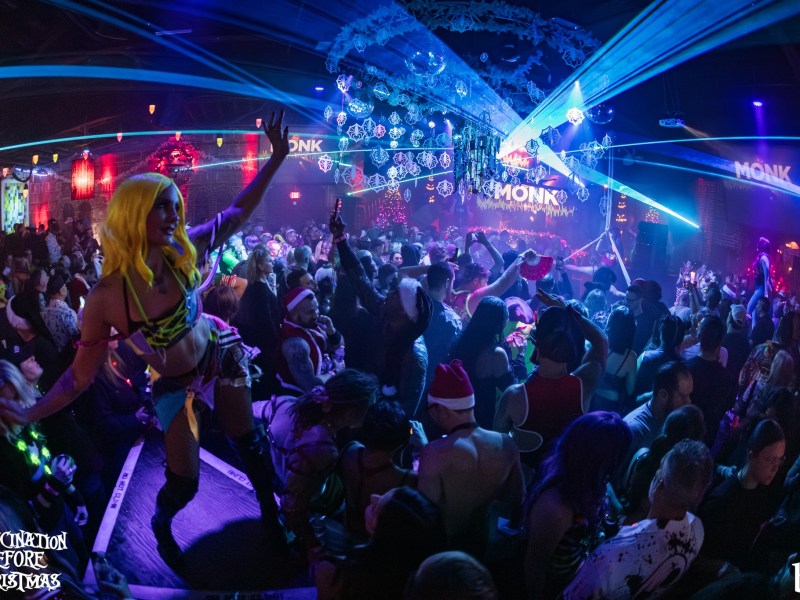 A large group of people celebrating at a club. A performer in the foreground, wearing a yellow wig and a brightly colored outfit, strikes a dramatic pose with a hand raised on a platform overlooking the crowd.