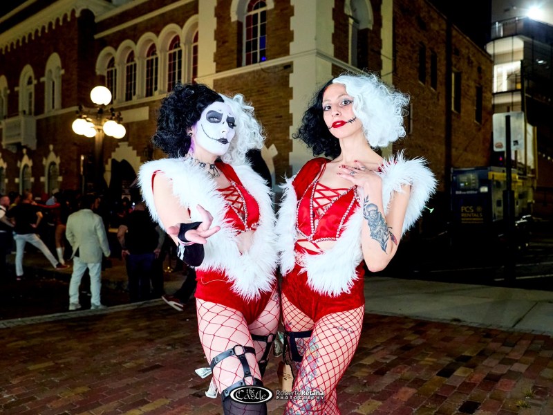 A promotional photo of two performers outdoors at night, standing in front of an historic red brick building. The subjects wear high-contrast outfits with black and white hair, red lips, and dramatic makeup.