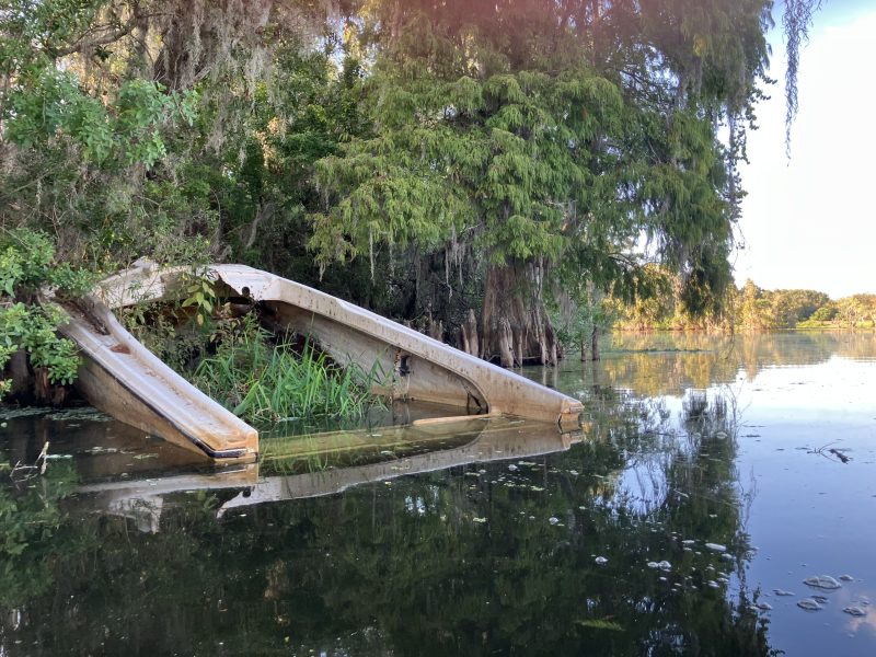 A dilapidated, abandoned white boat is partially submerged in murky swamp water near the tree-lined bank, surrounded by cypress trees and moss.