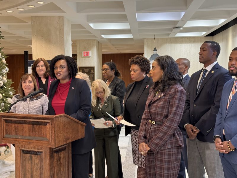 A diverse group of people, formally dressed, are gathered in a brightly lit, modern hall with white ceiling panels and columns. Several members stand to the right of the podium, and a decorated Christmas tree is visible to the left.