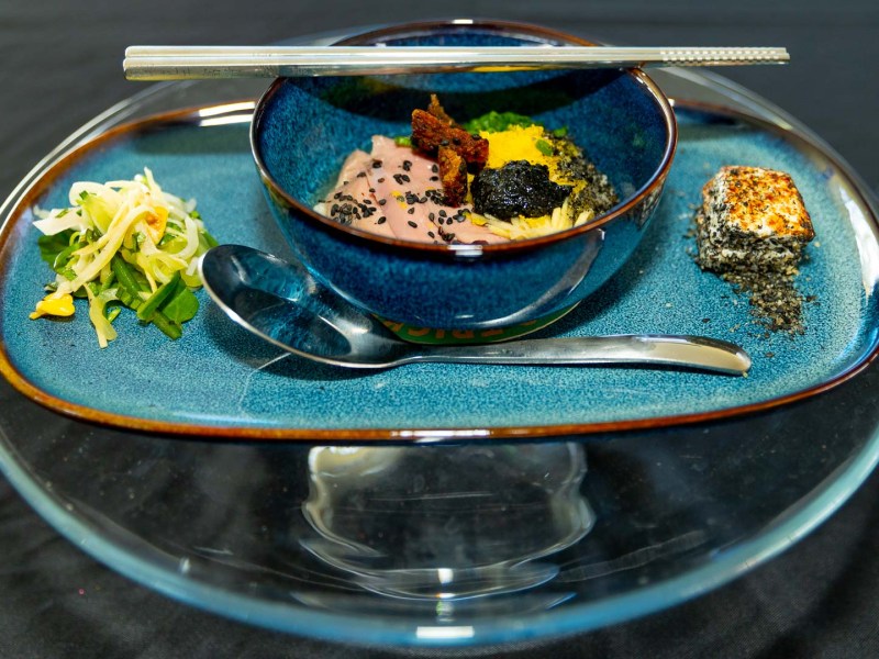 A close-up, high-angle shot of a gourmet dish. In the center, a deep blue bowl holds noodles, seared pink meat sprinkled with black sesame seeds, and colorful garnishes. Silver chopsticks lie across the bowl's rim. The bowl is on a large, blue-green speckled platter, which also holds a small side salad to the left and a sesame-seed-crusted block of food to the right. A silver spoon rests in front of the bowl on the platter.