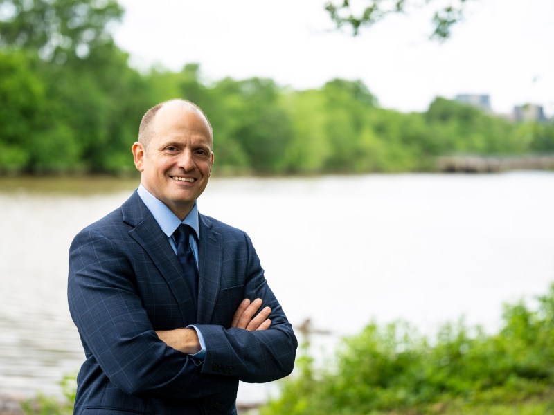 A portrait of Jose Javier Rodriguez, a man with a receding hairline, standing with his arms crossed. He is smiling warmly at the camera and wearing a blue pinstripe suit, a light blue shirt, and a tie. The background is a blurred, lush green park with a body of water behind him.