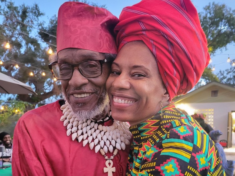 A close-up of a smiling older Black man and a woman embracing cheek-to-cheek at an outdoor gathering. The man wears a red hat, glasses, and a prominent cowrie shell necklace with a large Ankh pendant. The woman wears an elaborate, tall red headwrap and a vibrant top featuring African-inspired geometric patterns in yellow, green, and black. String lights glow in the background against a twilight sky.