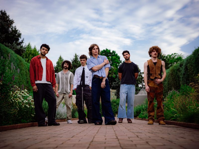 A low-angle, full-body outdoor shot of the six members of the band Ax and the Hatchetmen standing on a brick pathway. The group is positioned in front of lush, tall green hedges and some flowering plants under a cloudy sky. The members are wearing casual, individualistic clothing. From left to right: a man in a red and black plaid shirt over a white tee; a man in a light grey striped cardigan over a white shirt; a man in a white dress shirt with a black tie and black pants; the central figure, a tall man in a light blue short-sleeved button-up shirt and dark wide-leg jeans, striking a confident pose; a man in a black t-shirt and light wash jeans; and a man in a brown suede vest and matching trousers with a prominent mustache and curly hair.