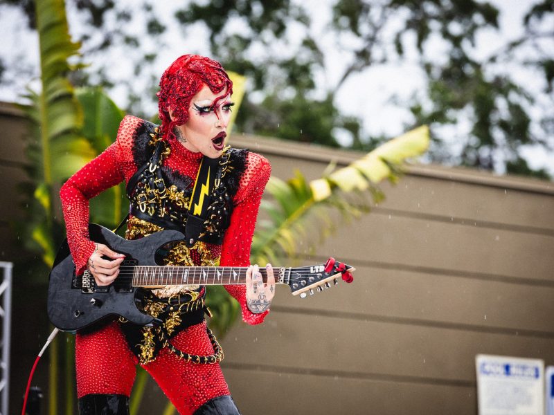 A performer with elaborate red hair and dramatic makeup plays a black electric guitar on an outdoor stage in the rain. They are wearing a red, crystal-encrusted bodysuit with gold detailing and a black harness featuring a lightning bolt strap. Raindrops are visible against the background of trees and a building, where a "POOL RULES" sign can be seen in the lower right corner.