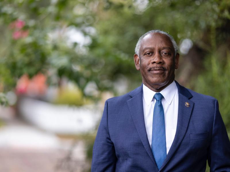 Outdoor portrait of Mayor Jerry Demings, wearing a dark blue suit and a patterned blue tie, smiling slightly against a blurred green, leafy background.