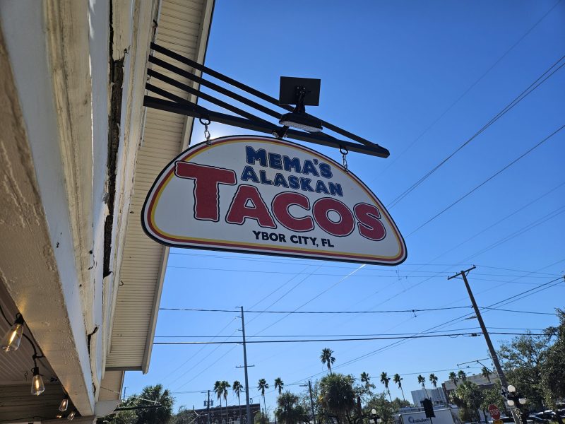 anging sign for Mema’s Alaskan Tacos in Ybor City, Florida, with bold red “TACOS” lettering. The sign is mounted on the side of a building with string lights underneath the overhang. Power lines, palm trees, and a clear blue sky are visible in the background.