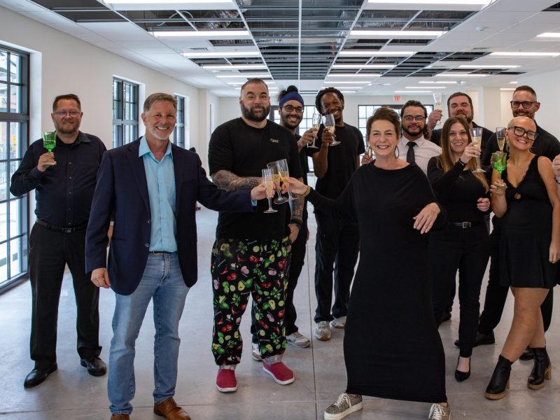 A large group of people celebrating inside a bright, new commercial space, raising glasses of champagne. The group includes a man in a blue blazer and a woman in a long black dress, surrounded by staff in black attire.