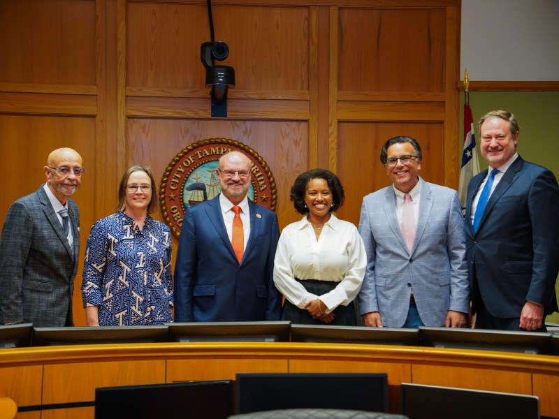 Group photo of six people, including a Black woman in a white blouse (Naya Young, center-right), standing in a city council chamber in front of the official City of Tampa seal and a wood-paneled wall.