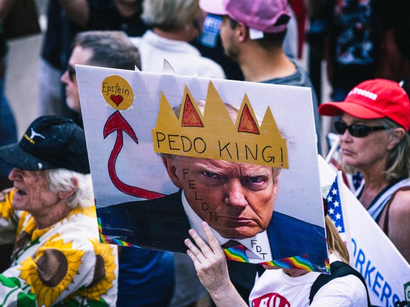 A close-up shot of a satirical protest sign being held up in a crowd. The sign superimposes a yellow crown reading "PEDO KING!" onto a photo of Donald Trump and includes a red arrow pointing to a heart labeled "Epstein." The crowd of diverse protesters is visible in the blurred background.