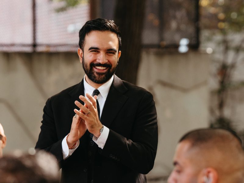 Portrait of a smiling man with a beard, wearing a black suit and tie, clapping his hands together outdoors at a campaign event.