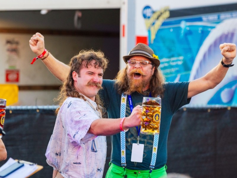 Two men celebrating at an outdoor festival. The man on the left has a mullet and a large mustache, wearing a white patterned shirt. The man on the right has a long red beard, is wearing a hat, suspenders, and green shorts, and is holding a large, full beer stein aloft. Both men have their free arms raised in excitement.