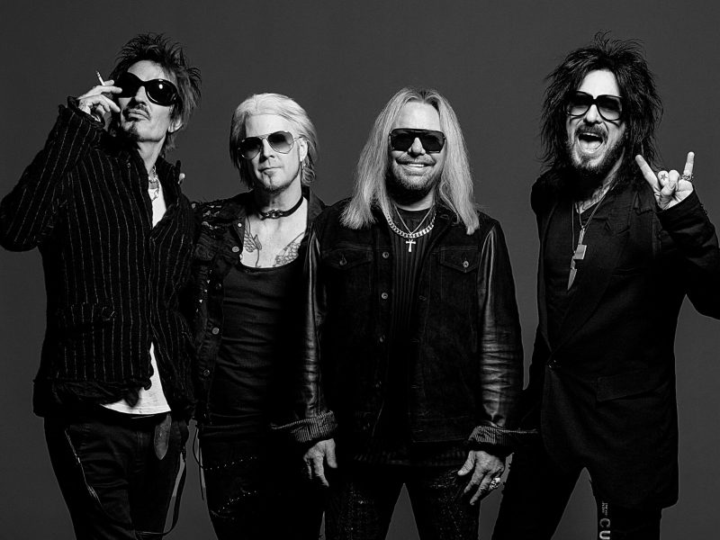 A publicity photo of Mötley Crüe in black and white. From left to right: Tommy Lee is smoking a cigarette; John 5, with light hair and a choker, stands next to him; Vince Neil is centered in a black leather jacket and sunglasses; and Nikki Sixx is on the far right, making a rock-on hand gesture.