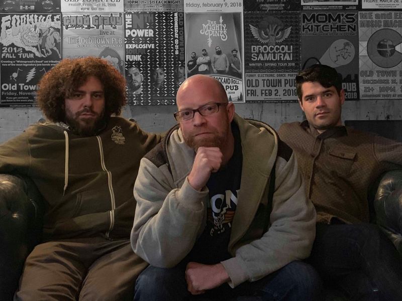 A trio of musicians. The person on the left has long, curly hair and a beard; the central person is leaning forward with a hand on their chin; and the person on the right looks directly at the camera.