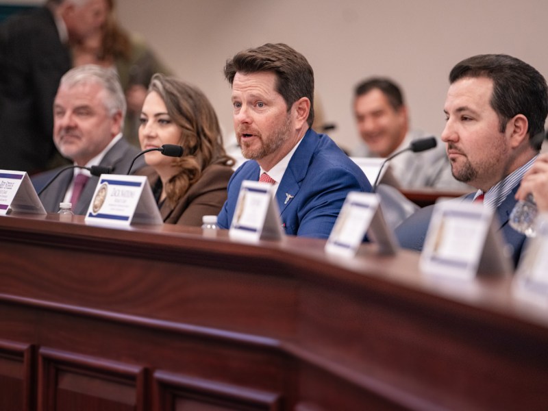 Representative Kevin M. Steele speaks at a legislative committee hearing, wearing a blue suit and red tie, seated at a long, dark wood desk with other members;