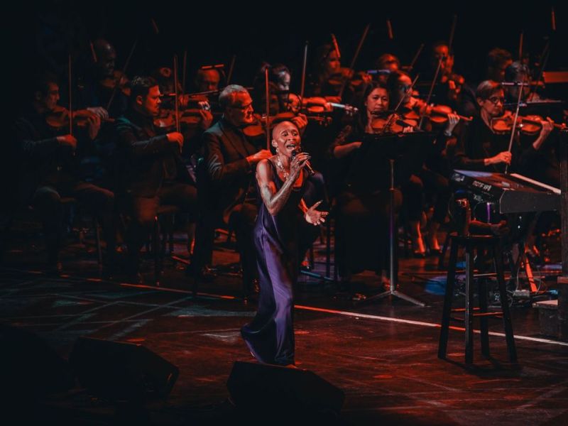 Singer Cynthia Erivo, dressed in a flowing purple gown, performs on stage with a microphone in hand, standing confidently in front of The Florida Orchestra.