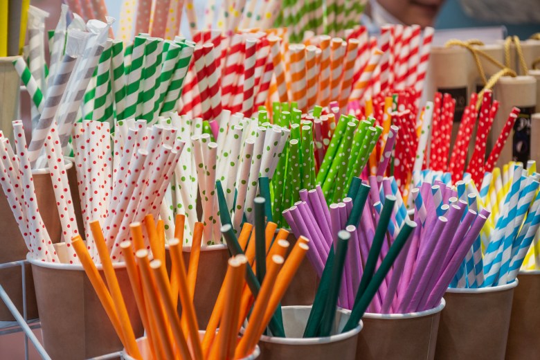 A close-up display of assorted colorful paper straws arranged in small brown cups. The straws feature a variety of vibrant patterns, including diagonal stripes and polka dots in red, green, blue, yellow, and purple, alongside solid-colored orange and purple straws.