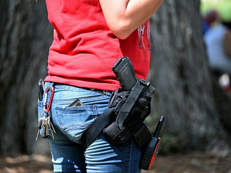 A close-up view from the waist down shows a person standing outdoors, wearing blue jeans and a red t-shirt. A black handgun is secured in a black thigh holster on their right leg, with a walkie-talkie clipped just below it. On the left side, a set of keys hangs from a red carabiner on a belt loop, and a card protrudes from the back pocket. The background is blurred, showing a large tree trunk and indistinct figures.